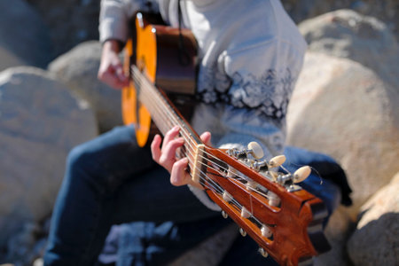 A performer of songs with a guitar. A woman plays the guitar, close-up, focus on the strings and neck of the guitar. The musician is sitting on large rocks in the mountains of Kyrgyzstan. The guitaristの写真素材