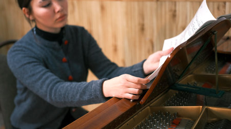 A serious young Asian woman is sitting at the piano and reading sheet music. She's wearing a gray sweater and she's focused on the music. Music school. A piano student learns to play the piano and learns a new melody. Inspirationの写真素材