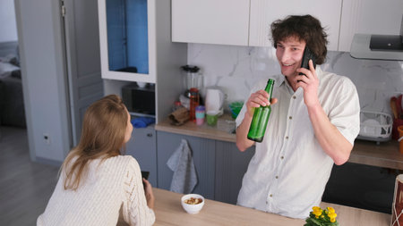 A happy man is talking on a cell phone with a bottle of beer in his hand. He has a girlfriend visiting. The beginning of the party. The relationship between the sexes.の写真素材