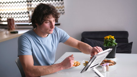 A single man is having breakfast in front of a tablet monitor. He's wearing a blue shirt, and there's a bowl of food in front of him. A student is sitting in the kitchenの写真素材