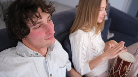 A young woman plays the drum for her boyfriend. A handsome man listens attentively to her rhythm. The students are sitting on the couch having fun. The woman is smiling and enjoying the music. The concept of relaxation and enjoymentの写真素材