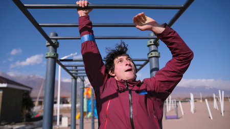 An athletic guy is training on a horizontal bar outside. A man in a red jacket is hanging from a...