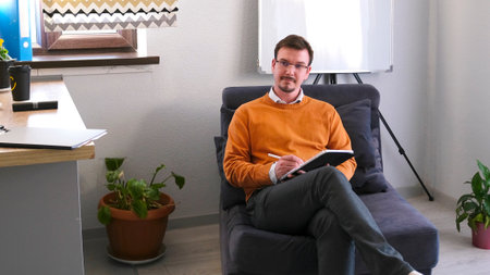 Psychologist's office. A young man with glasses is sitting on a blue armchair with a notebook and pen in his hands. He's wearing an orange sweater and he's focused on writing. The room has a kitchen area with a counter and a potted plant.の写真素材