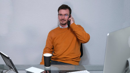A businessman is sitting at an office desk and talking on a mobile phone. There is a computer...