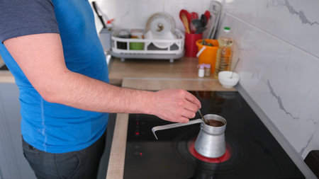 A man makes coffee in a Turk in the kitchen. He uses a metal coffee pot and a spoon to stir the coffee. The kitchen is well equipped with various utensils and household appliances, including a sink and refrigerator.の写真素材