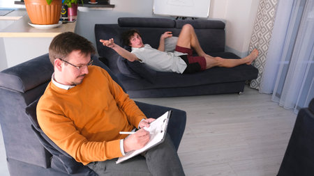 A successful psychologist in an orange sweater writes something on a tablet, while a young man lies on the couch and tells him about his problems. A man gives advice to a young manの写真素材