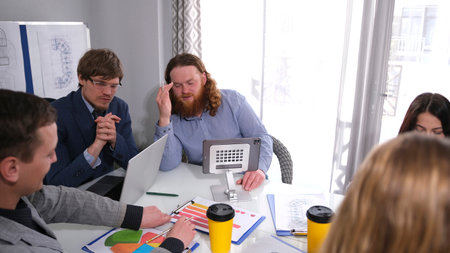 A handsome red-haired man is the boss's personal assistant, who gives him important information. A group of people are sitting at a table with laptops and cups of takeaway coffee.の写真素材