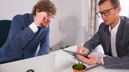 Two young entrepreneurs with glasses are sitting at a table, one of them is writing something on a...