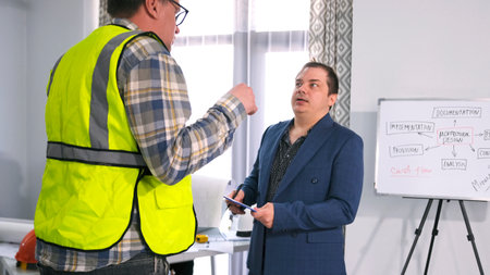 A male engineer in glasses and a green signal vest is talking to the head of the company. Two men are discussing the project estimate. In the office of the architectural bureau.の写真素材
