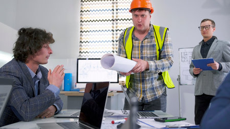 A male foreman in a green signal vest and an orange construction helmet holds a piece of paper and...