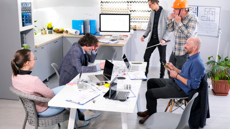 A worker puts on an orange construction helmet. A group of architects and engineers are sitting at a...