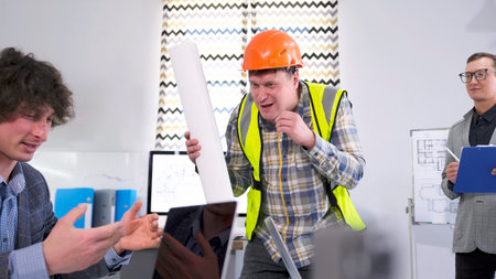 A contractor in a green vest is laughing and wearing an orange hard hat, holding up blueprints of the building and arguing with the company's manager. Meeting at the architectural bureau.の写真素材