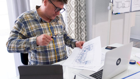 A man with glasses is sitting at a table with a laptop and a stack of blueprints. He holds a...