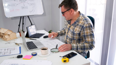 A guy in glasses and a plaid shirt is sitting at a desk with drawings, a laptop and a tablet. He's...