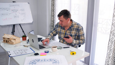 The designer is sitting at a desk with a laptop, building drawings and a mobile phone. He is focused...