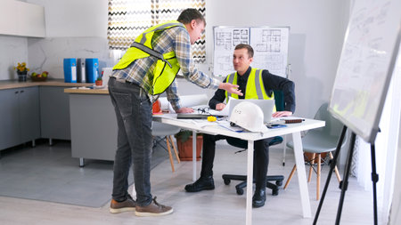 Architectural office. Two male workers in green vests are talking about working on a project. The...