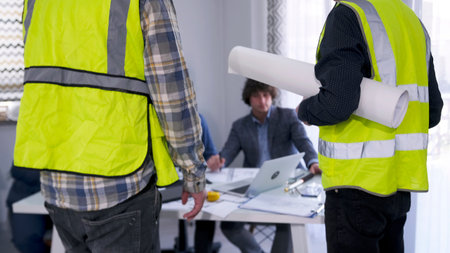 A foreman and a worker in green vests with drawings in their hands stand at the desk of the head of...