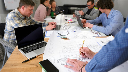 Close-up of the architect's hands making changes to the drawings. A group of people is working...
