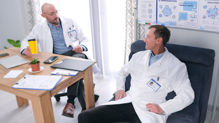 A young, handsome intern is chatting with his older friend. Two men in white lab coats are sitting...