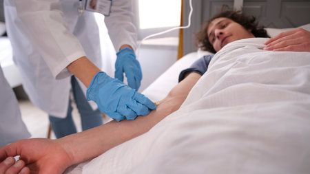 A curly-haired student is lying on a hospital bed. An intern stands next to him and puts a saline drip on the patient. A doctor in a white lab coat and blue medical gloves performs a standard procedure at the clinic.の写真素材