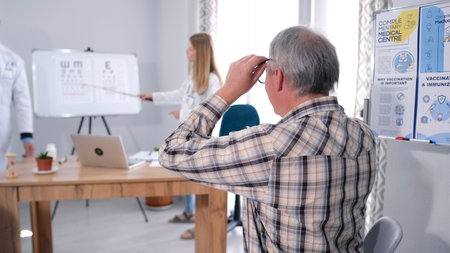 A gray-haired man in a plaid shirt is sitting in an ophthalmologist's office and checking his eyesight. A young intern woman stands at a blackboard with vision test tables and points to one of the letters.の写真素材