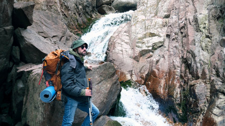 A lonely man in a hat, sunglasses, with a backpack and trekking poles stands in front of a waterfall...