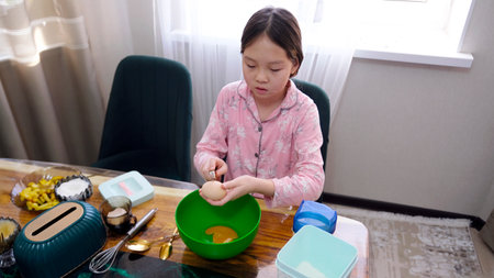 A young Asian girl cooks pancakes on the kitchen table in the early morning. A child in pink pajamas...