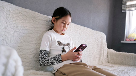 A young girl is sitting on the couch and looking at her mobile phone. An Asian child is intently...