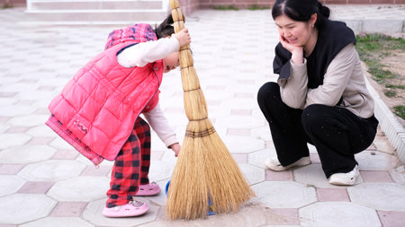 Sweep the floor. A woman is kneeling next to a child who is holding a broom and a dustpan. Mom lovingly watches as her little daughter helps her with the housework.の写真素材