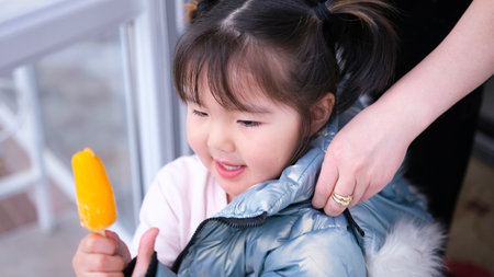 Close-up portrait of a happy Asian girl with a popsicle in her hand. Mom helps put a jacket on her little daughter while she eats ice cream.の写真素材