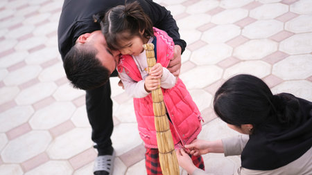 Dad kisses his little daughter, and mom buttons her jacket. An Asian man hugs a girl, and a woman...