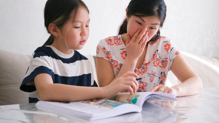 Mom helps her daughter do her homework. A woman and a child are sitting at a table and reading a school textbook to complete homework assignments. The familyの写真素材
