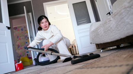 The housekeeper cleans the house. A woman cleans a room with a vacuum cleaner. She's focused on her task.の写真素材
