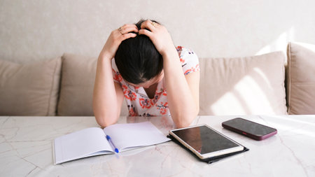 An anxious woman is sitting at a table with a notebook, phone and tablet. The tired woman hung her...