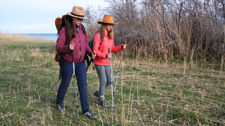 Two women went hiking along the lake. Wanderers are strolling through the field with backpacks and trekking poles. Hikers enjoy spending time outdoors.の写真素材