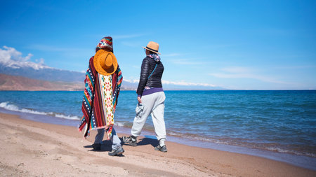 Two women in authentic outfits are strolling along the beach against the background of the blue...
