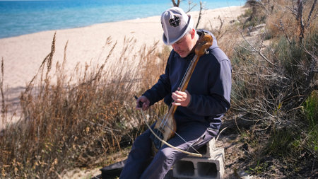 A lonely, thoughtful man is sitting on a wild beach with a national Kyrgyz musical instrument in his hands. Komuz or Kyl Kayak. The beach is visible in the background.の写真素材