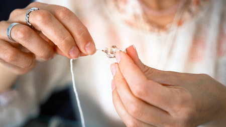 A woman holds a thread and a small shell in her hands, close-up. The needlewoman makes a necklace or...