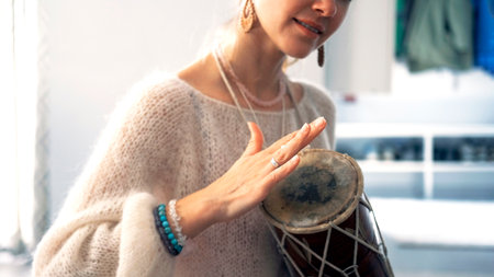 Close-up of a woman in a white knitted sweater playing a small drum. She is smiling and enjoying what is happening. Music and creativity. Hobbies and artの写真素材