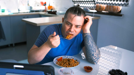 A man is eating in the kitchen. A guy eats fried bulgur with vegetables while watching a movie on his tablet. An ordinary day for a person.の写真素材