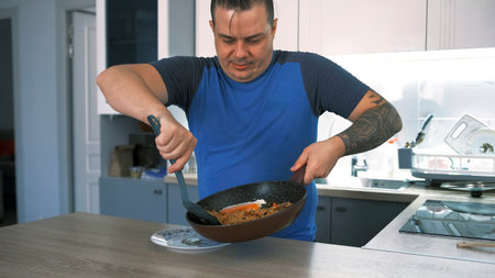 A guy has cooked himself a meal and is putting hot food from a frying pan on his plate. A male cook in the kitchen. He's wearing a blue shirt and has tattoos on his arm.の写真素材