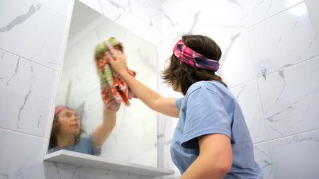 A woman wipes the bathroom mirror with a towel. The maid cleans the bathroom. The cleaner cleans the mirror. She's wearing a blue shirt and a pink headband.の写真素材
