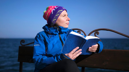 A woman is sitting on a bench by the sea and reading a book. She's wearing a blue jacket and a...