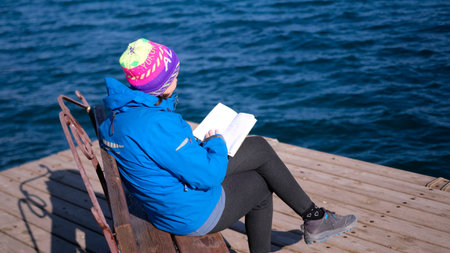A woman sits on a bench on a pier by the sea and reads a book. Literature and education. The student...