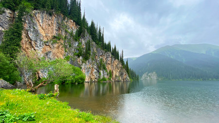 View of a sheer cliff covered with green fir trees near the Middle Lake Kolsai. A mountain range with a lake in the foreground. The sky is covered with clouds. Trekkingの写真素材