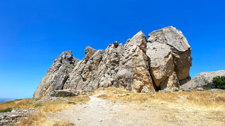 A large rock formation is located on a hillside with a clear blue sky above it. Beshbarmag is a mountain in Azerbaijan. The rocks are jagged and unevenの写真素材