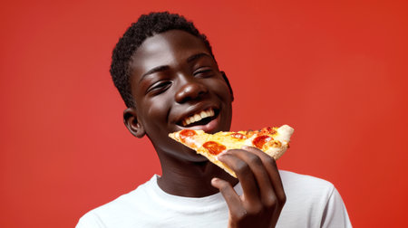 A happy black teenager in a white T-shirt holds in his hand a piece of pizza with pepperoni and cheese on a red background. He smiles and takes a bite of pizza. Italian cuisine. Pizza Dayの素材