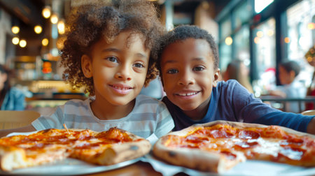 Two cute black kids are sitting in a pizzeria eating pizza. The little brother and sister smile as they look at two large pizzas. Italian cuisine. Pizza day. The scene is fun and playfulの素材