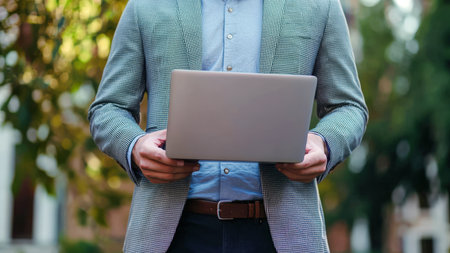 A stylish man with a laptop in his hands. The businessman is in a hurry on business. The concept of modern technologies. A guy walks down a street with green treesの素材