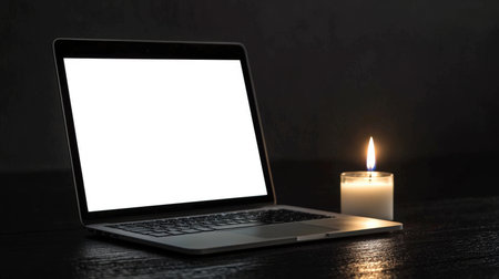 A laptop with a white screen stands in a dark room with a burning candle on the table. Laptop mockup. The concept of Earth Hour. Saving electricity. The stage creates a cozy and intimate atmosphere. The laptop is open and the screen is blank.の素材
